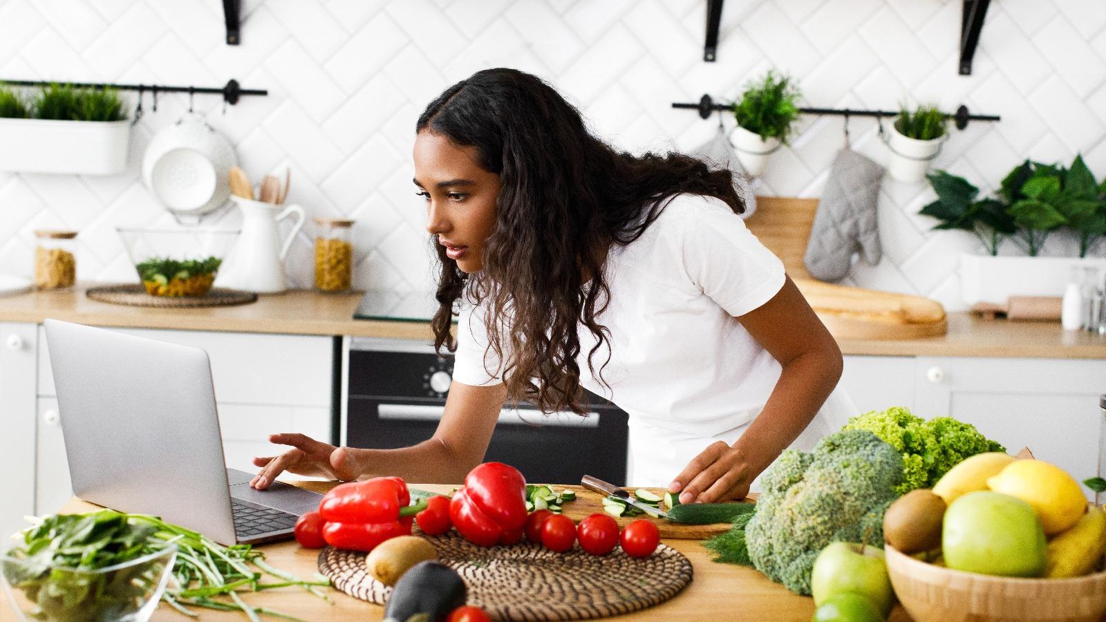 Young woman in kitchen looking at laptop while cooking