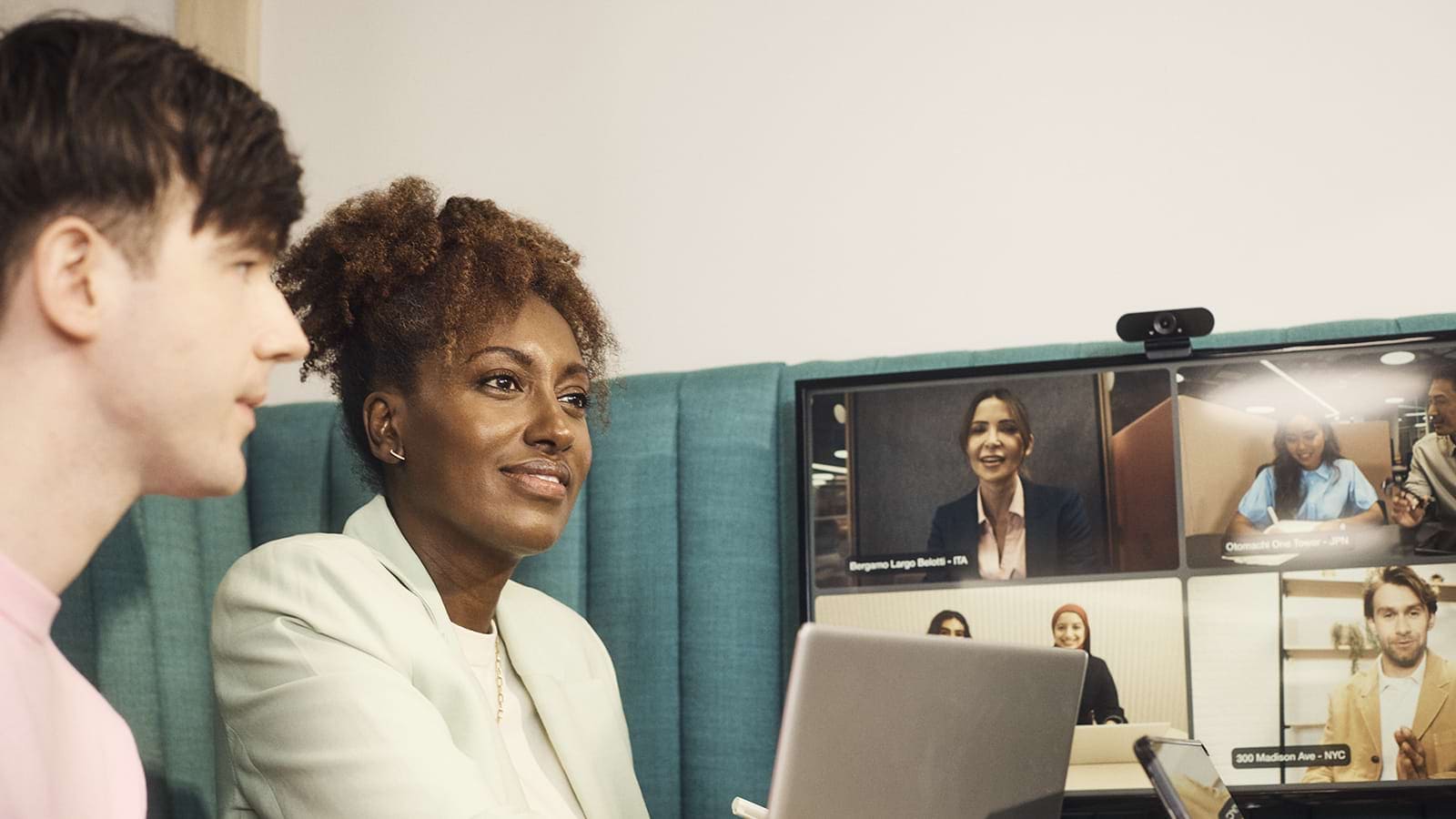 Two individuals are seated at a table with laptops, engaging in a virtual meeting displayed on a screen