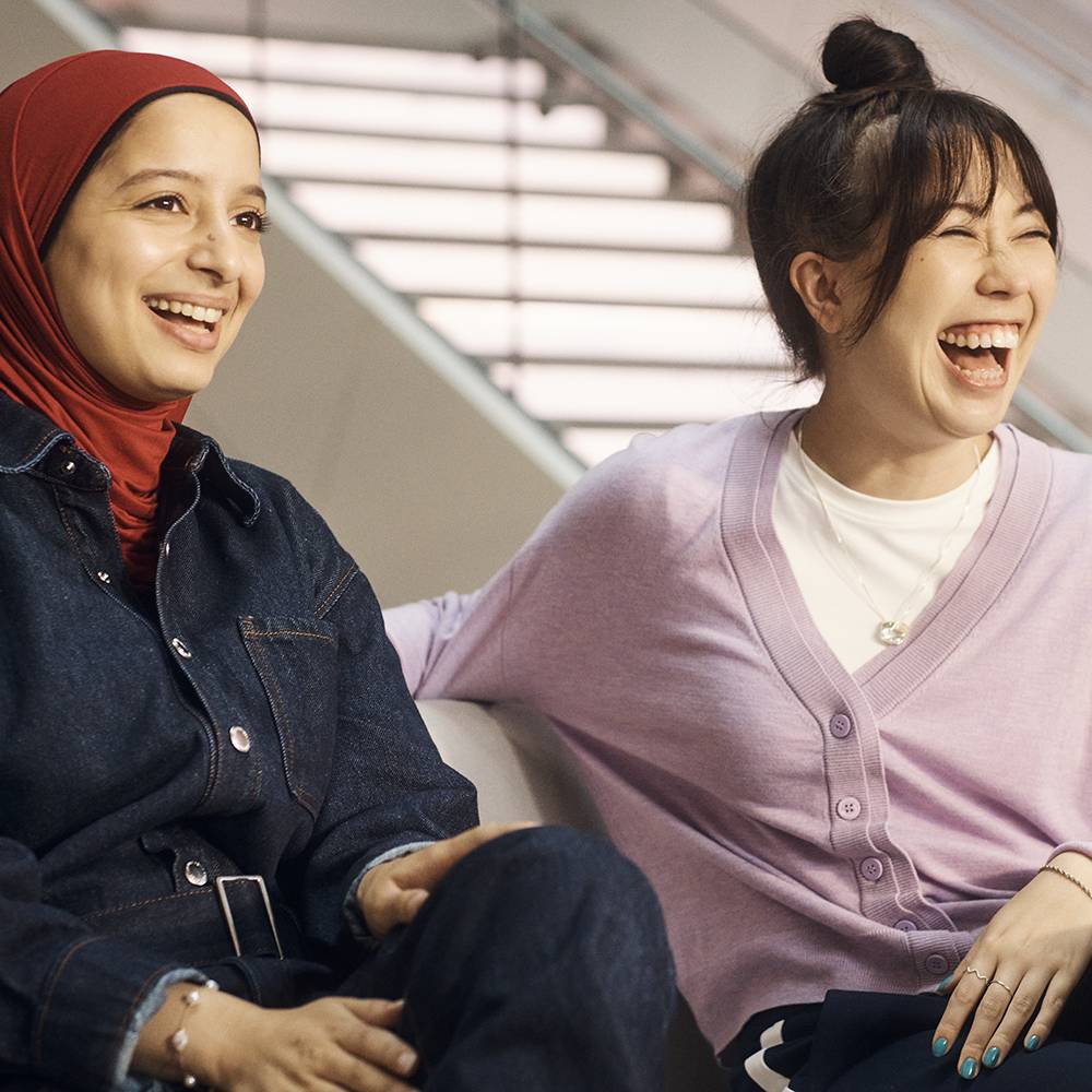 Two women are seated on a couch in a contemporary indoor environment with a staircase and plants visible in the background