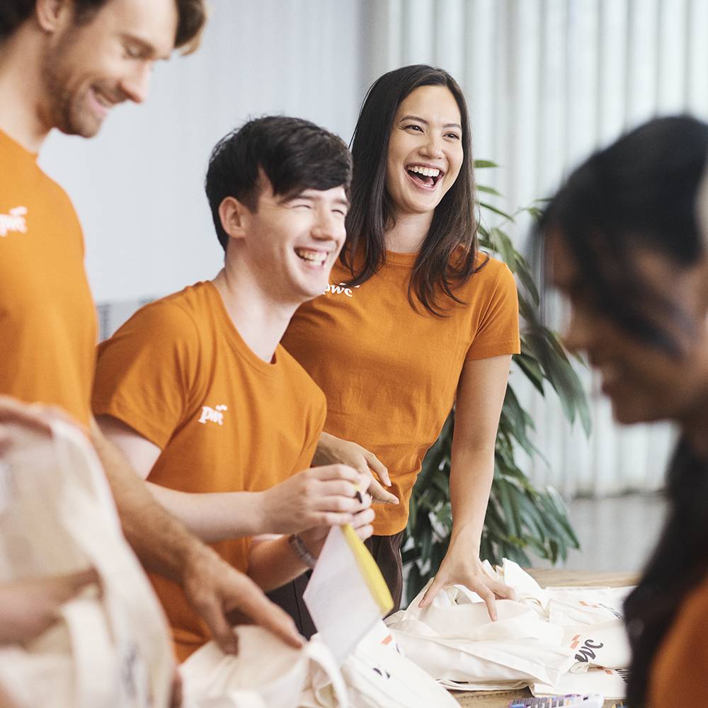 A group of individuals wearing orange PwC shirts assembling tote bags at a community event